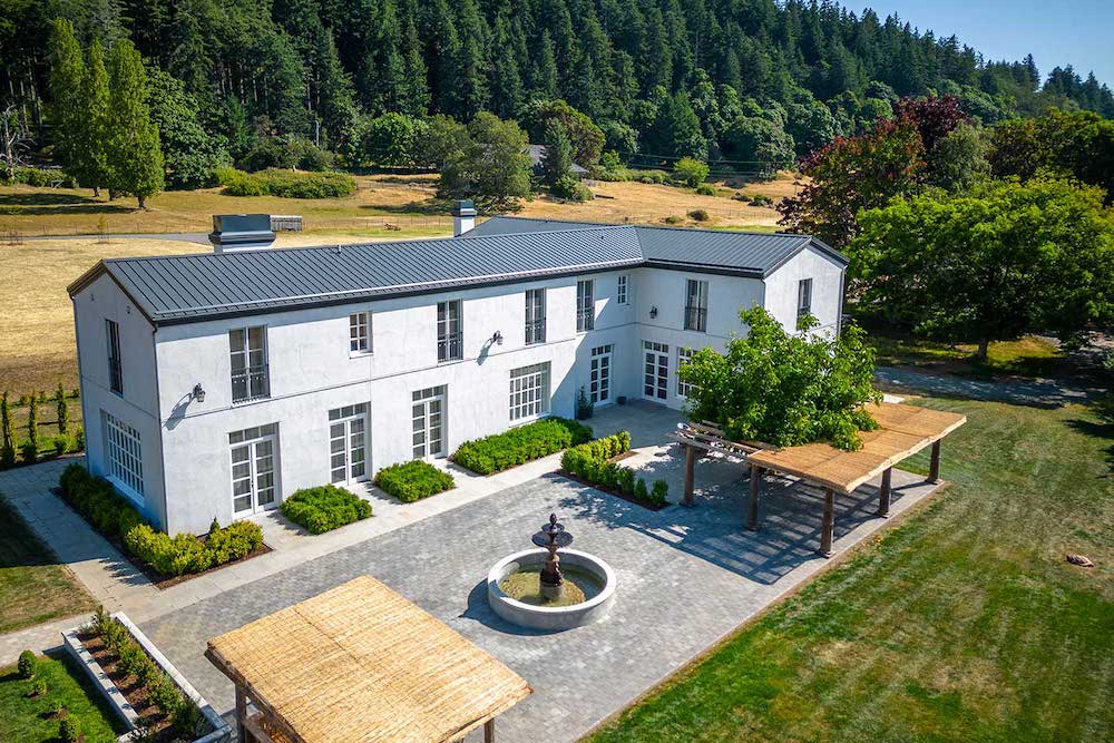 Elegant two-story coastal European-style building with a central fountain, pergola seating, and forest backdrop